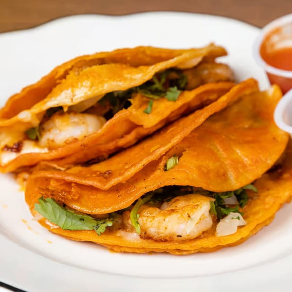 Three shrimp tacos with cilantro and onions on orange tortillas, served with two small sauce cups.