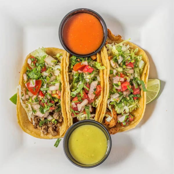 Three vibrant tacos with various fillings, lettuce, and pico de gallo, served with red and green sauces.