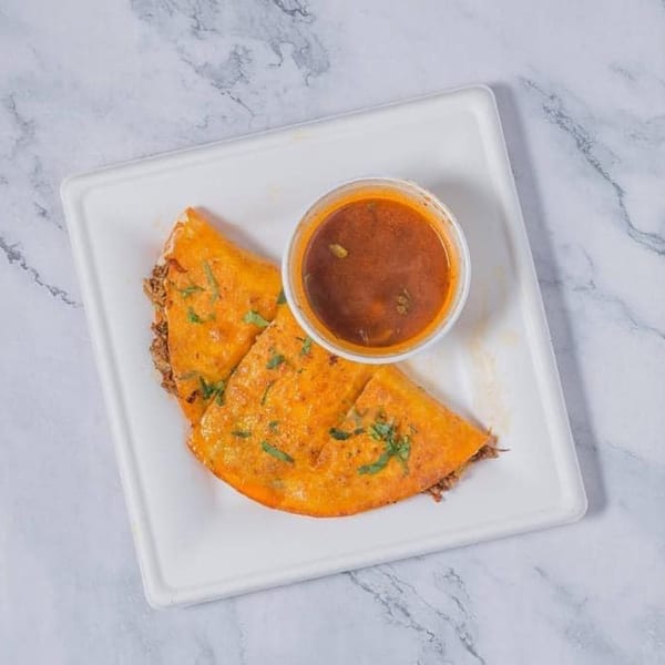 Two crispy birria tacos with cilantro on a white plate, served with a side of consommé.