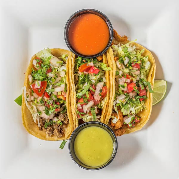 Three vibrant tacos with various fillings, fresh toppings, and two dipping sauces on a white plate.